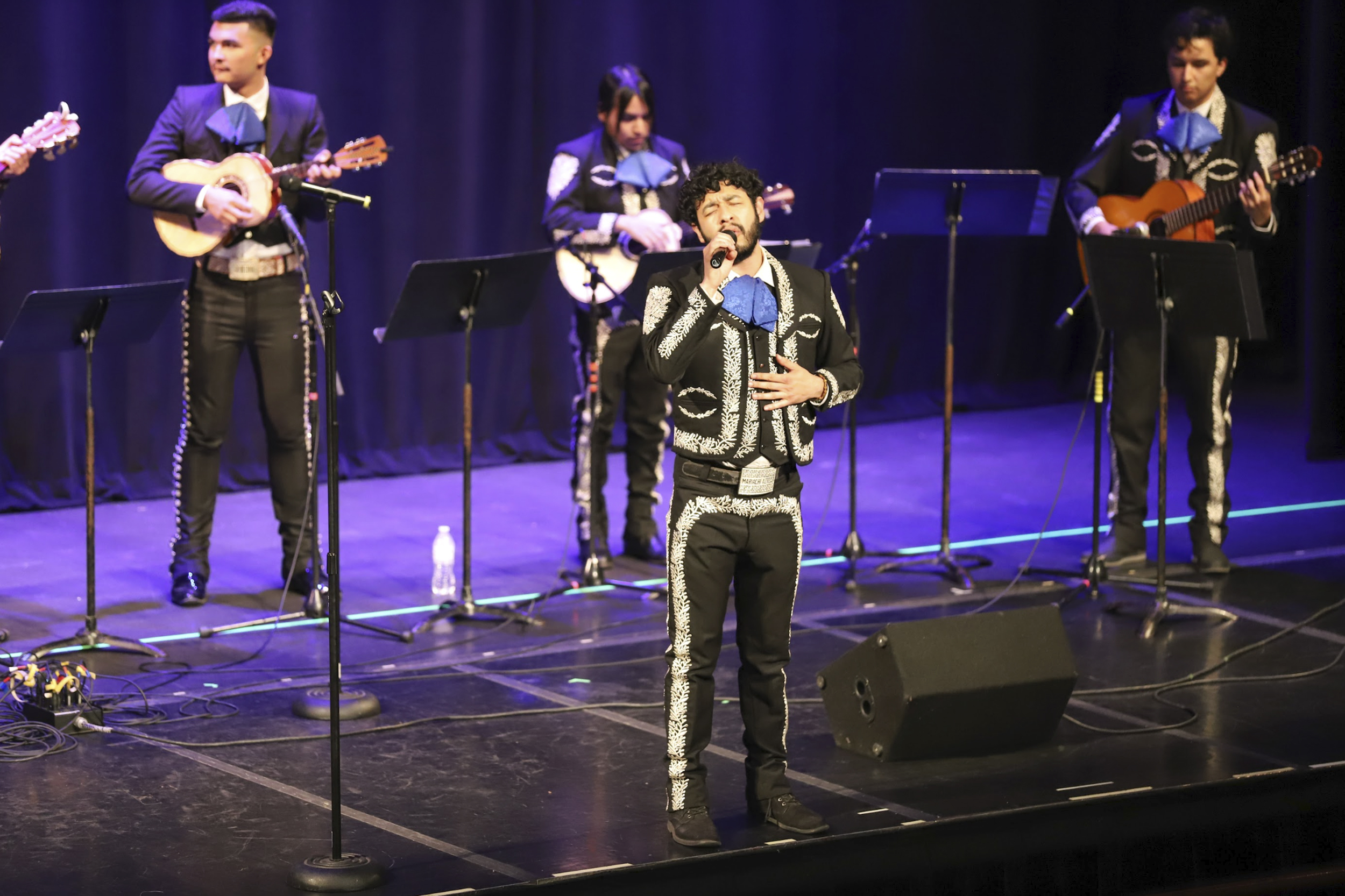 Photo of a mariachi ensemble performing. A dark-haired man sings passionately in the foreground.