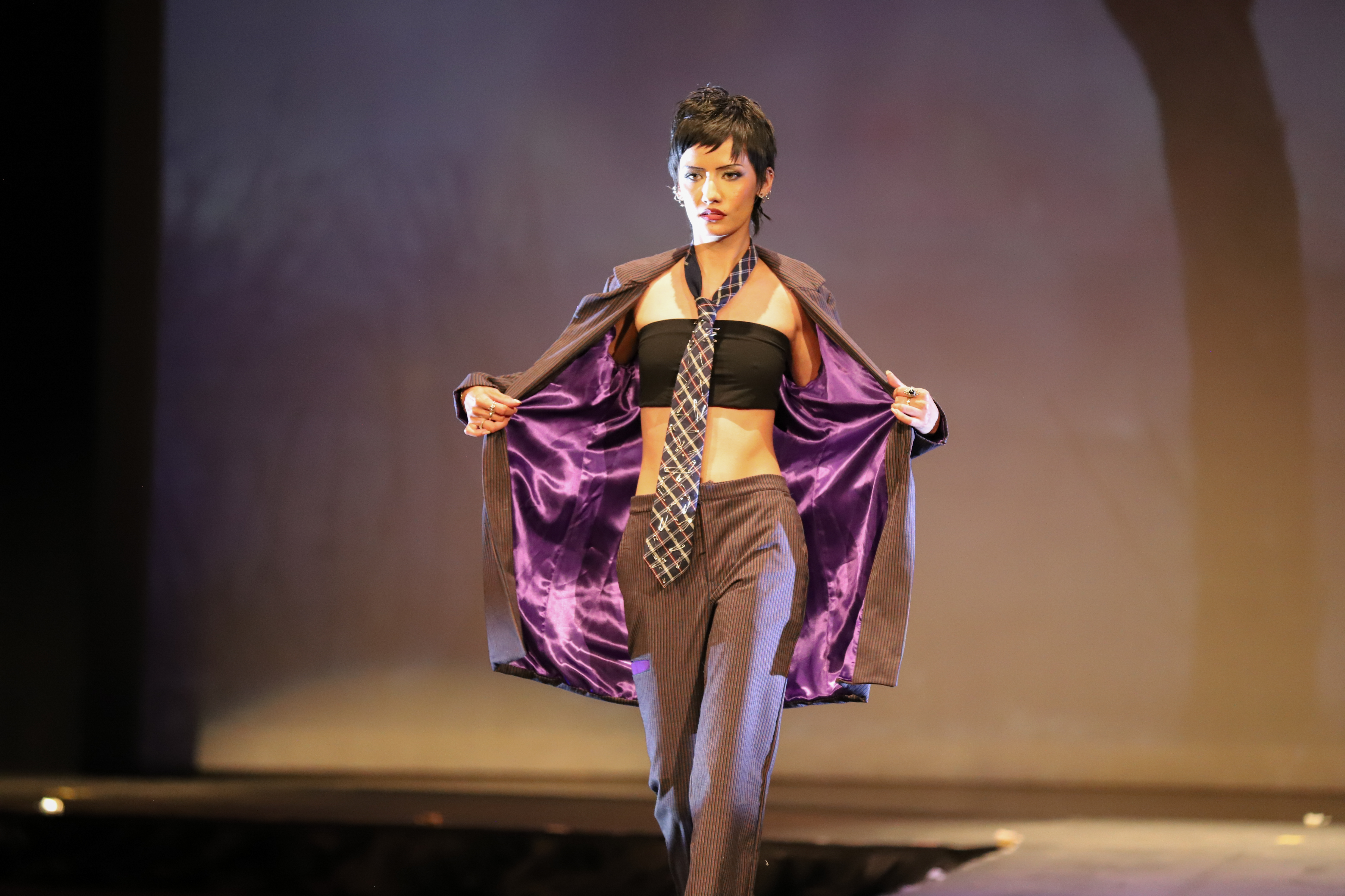 Photo of a runway model wearing a tie, bandeau top, and suit, holding open the jacket to reveal the purple lining.