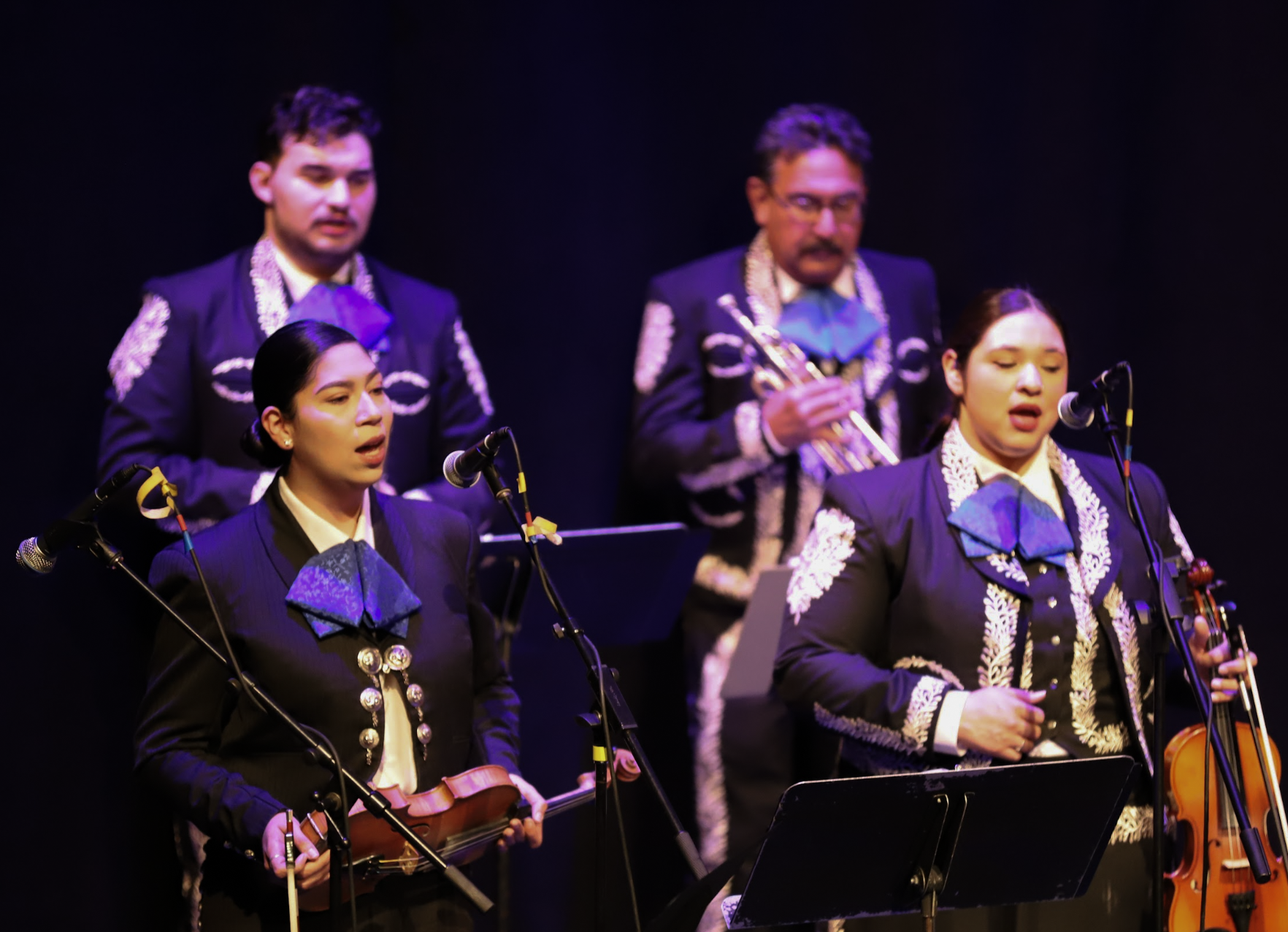 Photo of a mariachi band performing. Two woman violinists hold their instruments while they sing with other musicians behind them.