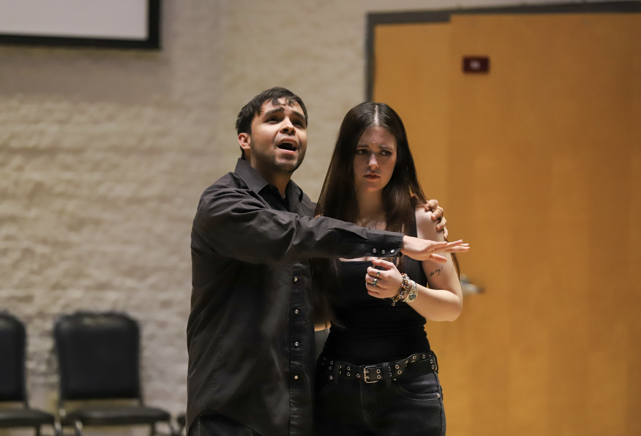 A young Latino man sings a show tune passionately with his arm around a long-haired woman, who looks distraught.