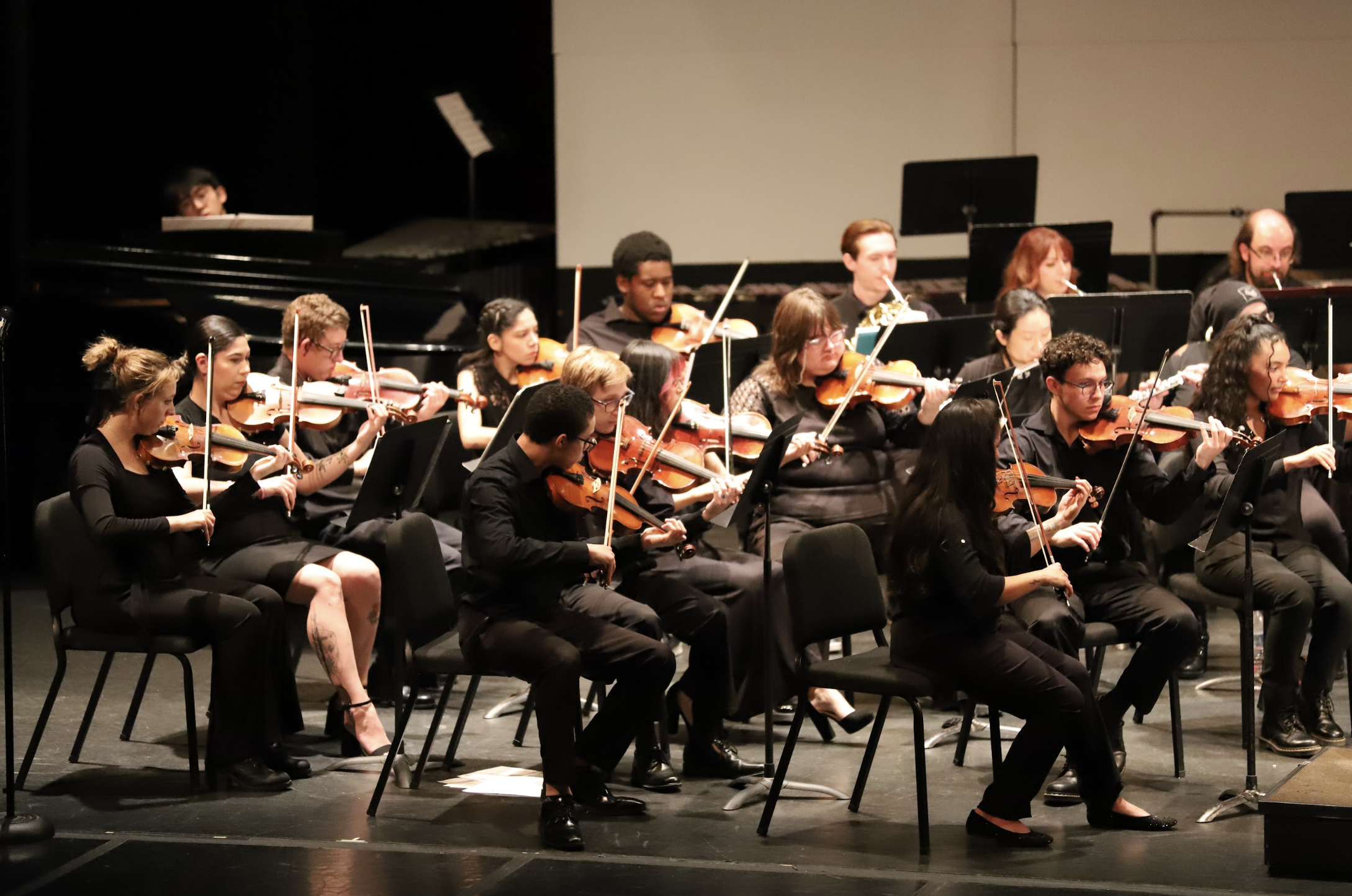 Photo of men and women violinists performing with the PCC Symphonic Orchestra. Behind them can be seen a man playing the piano and three French horn performers.