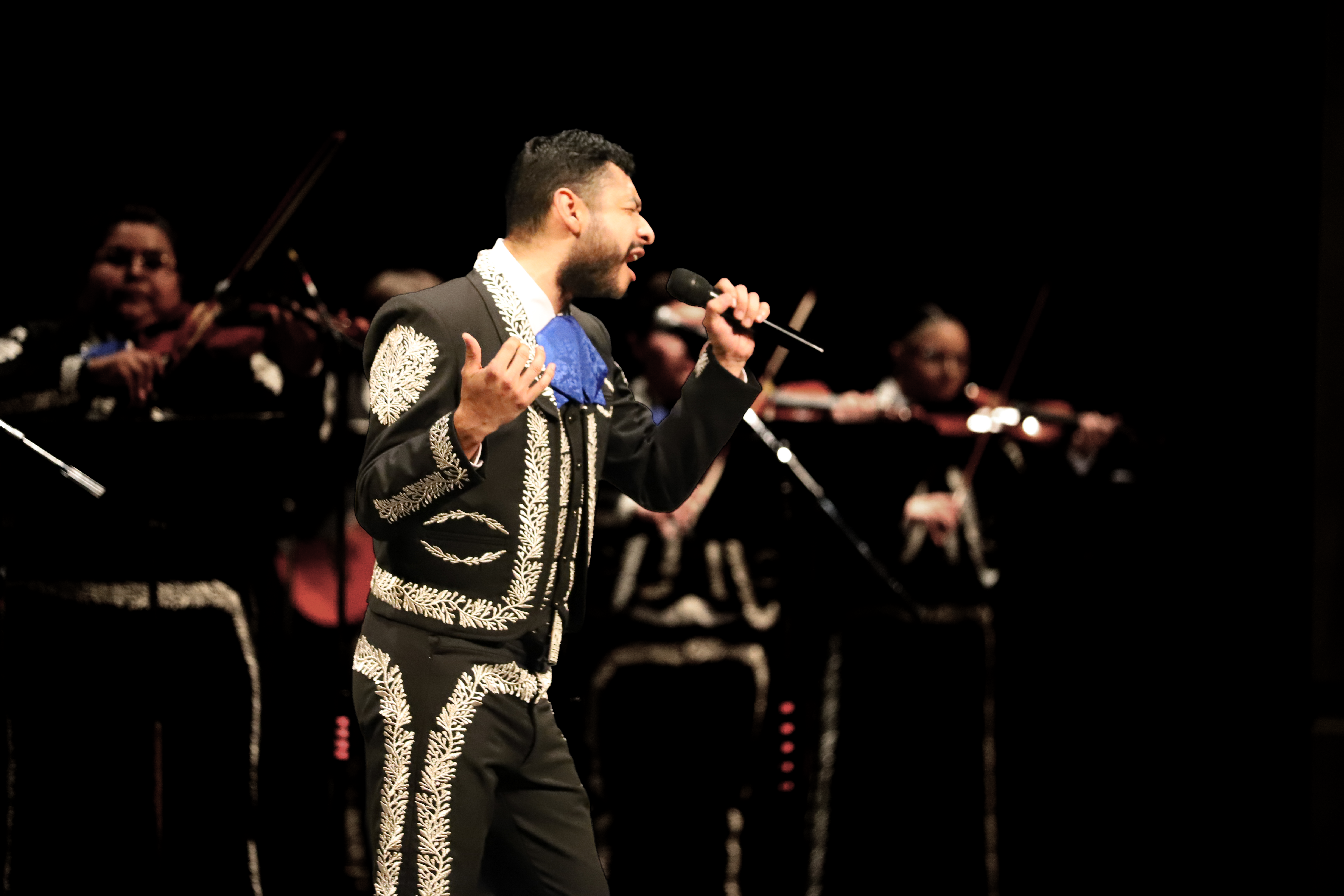 Photo of a mariachi band playing. In the foreground, a male performer sings passionately.